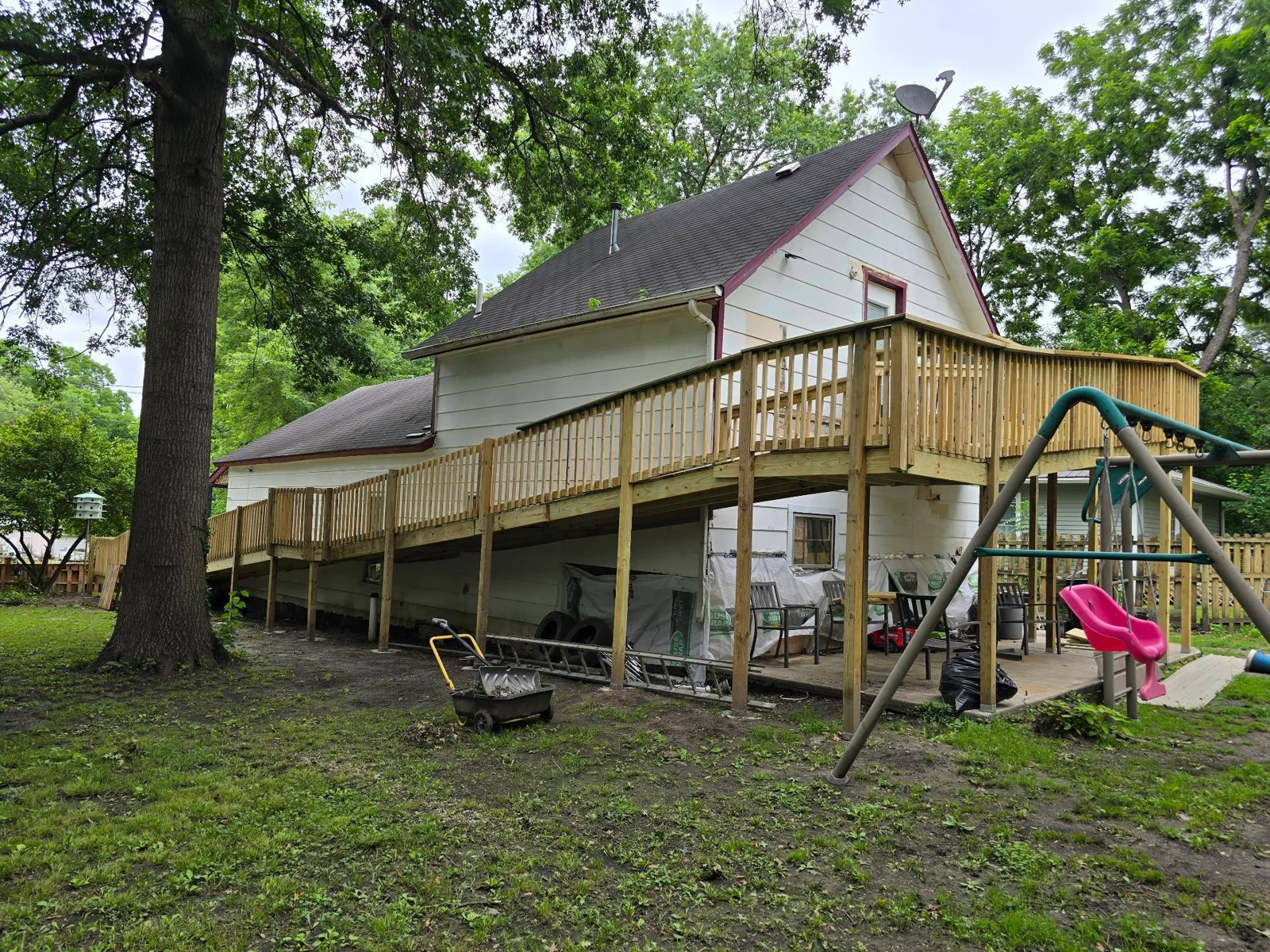 Side and rear view of a long wooden accessible ramp and deck beside a white house, with a large tree and swing set in the yard